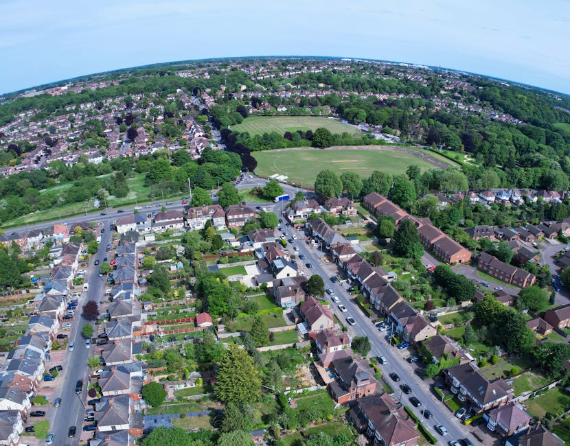 An aerial view of a residential suburb with rows of terraced houses and semi-detached homes, each with small, well-maintained gardens and lawns. The streets are lined with parked cars, and mature tree
